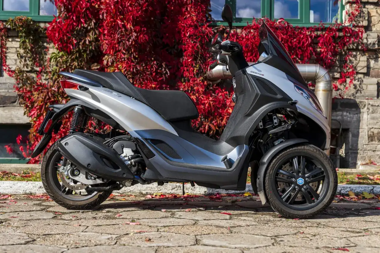 Srebrny Piaggio MP3 300, tr&oacute;jkołowy skuter, prezentuje swoje dane techniczne na tle jesiennych liści i ceglanej ściany.