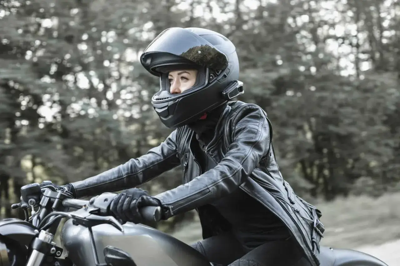 Kobieta w skórzanej kurtce i kasku szczękowym lub integralnym jedzie motocyklem przez las.