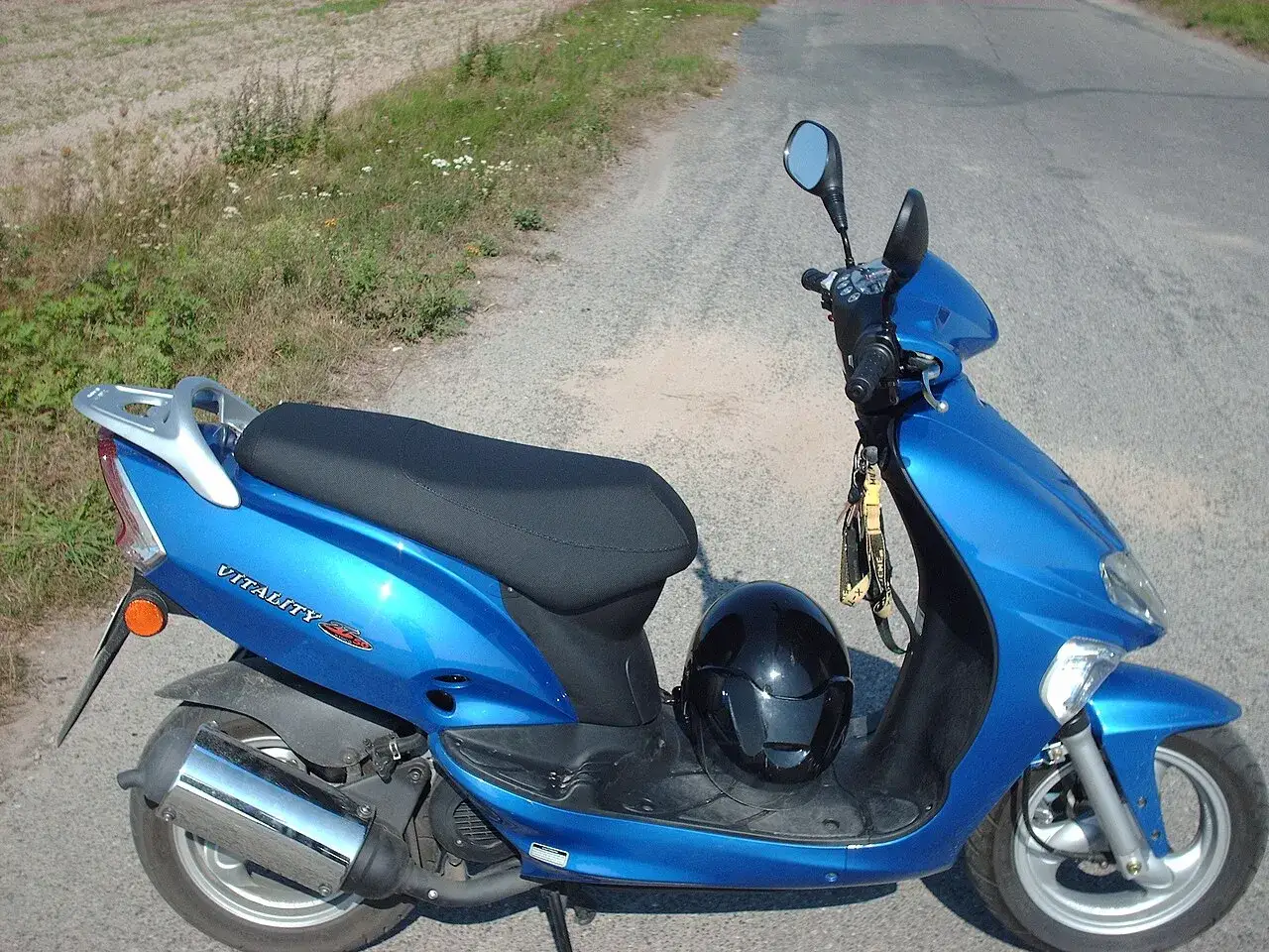 Niebieski skuter Kymco Vitality 50 2T z czarnym kaskiem na siedzeniu, zaparkowany na poboczu drogi.