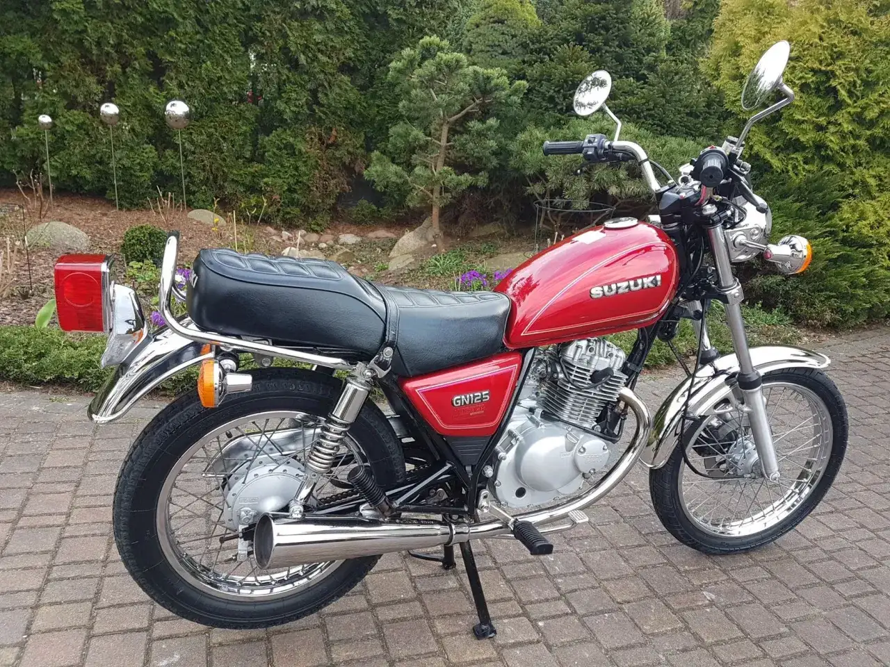 Czerwony motocykl Suzuki GN 125 z chromowanymi elementami, zaparkowany na brukowanej ścieżce wśród zieleni.