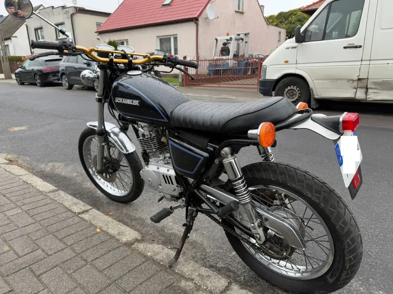 Czarny Suzuki GN 125 w wersji Scrambler, z błyszczącymi chromowanymi elementami i złotą kierownicą, stoi na chodniku.