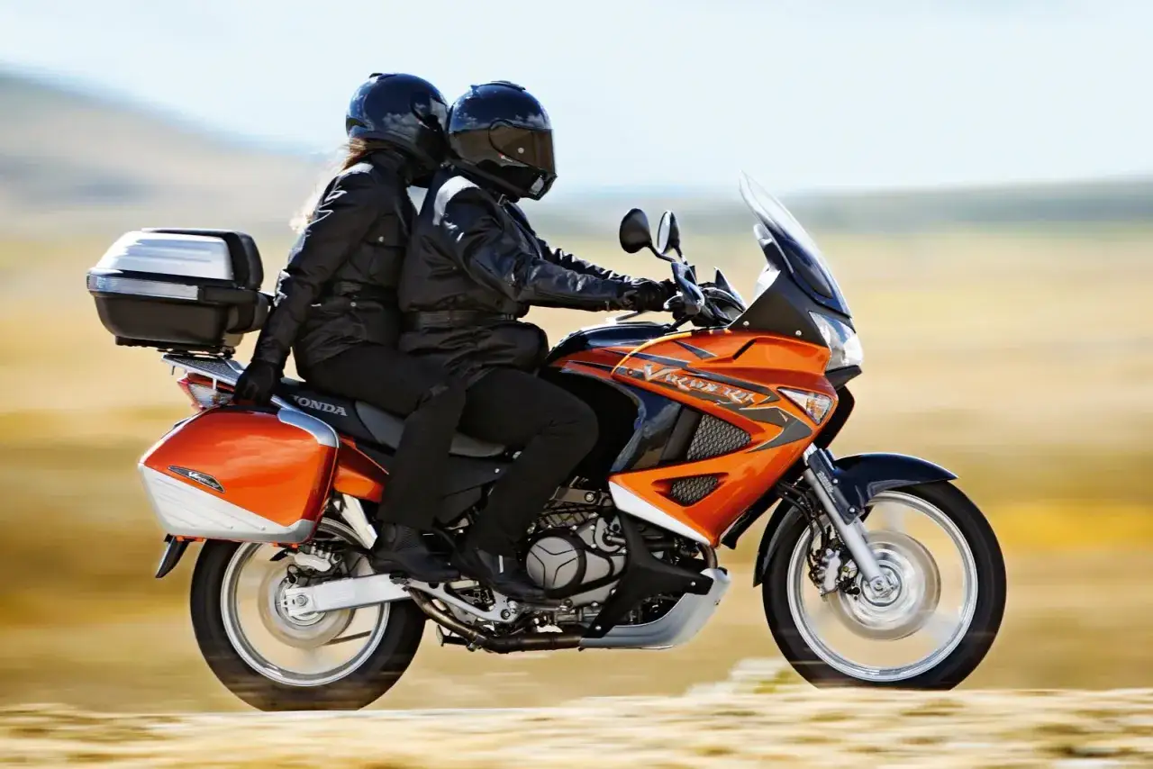 Pomarańczowa Honda Varadero XL 1000 z dwoma motocyklistami w kaskach, gotowa na przygodę.