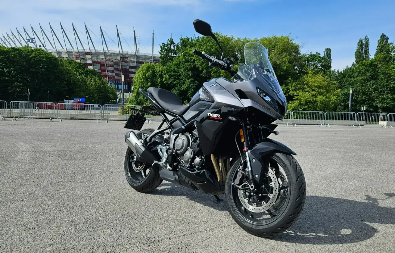 Szary motocykl Triumph Tiger Sport przed stadionem. Dane techniczne tego modelu są imponujące.