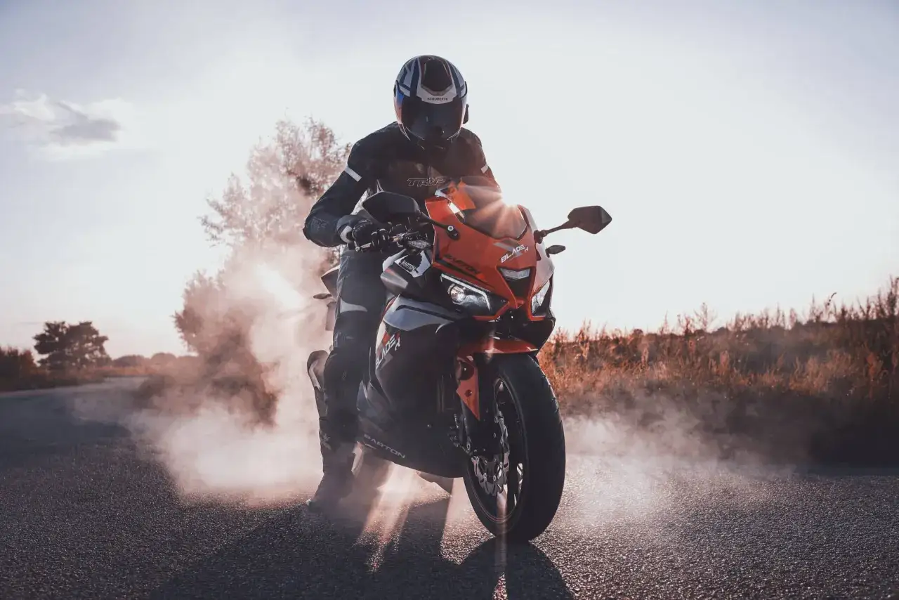 Motocyklista w kasku i kombinezonie na pomarańczowym Barton Blade 125, z którego unosi się dym. Zastanawiasz się, ile pali motor 125?