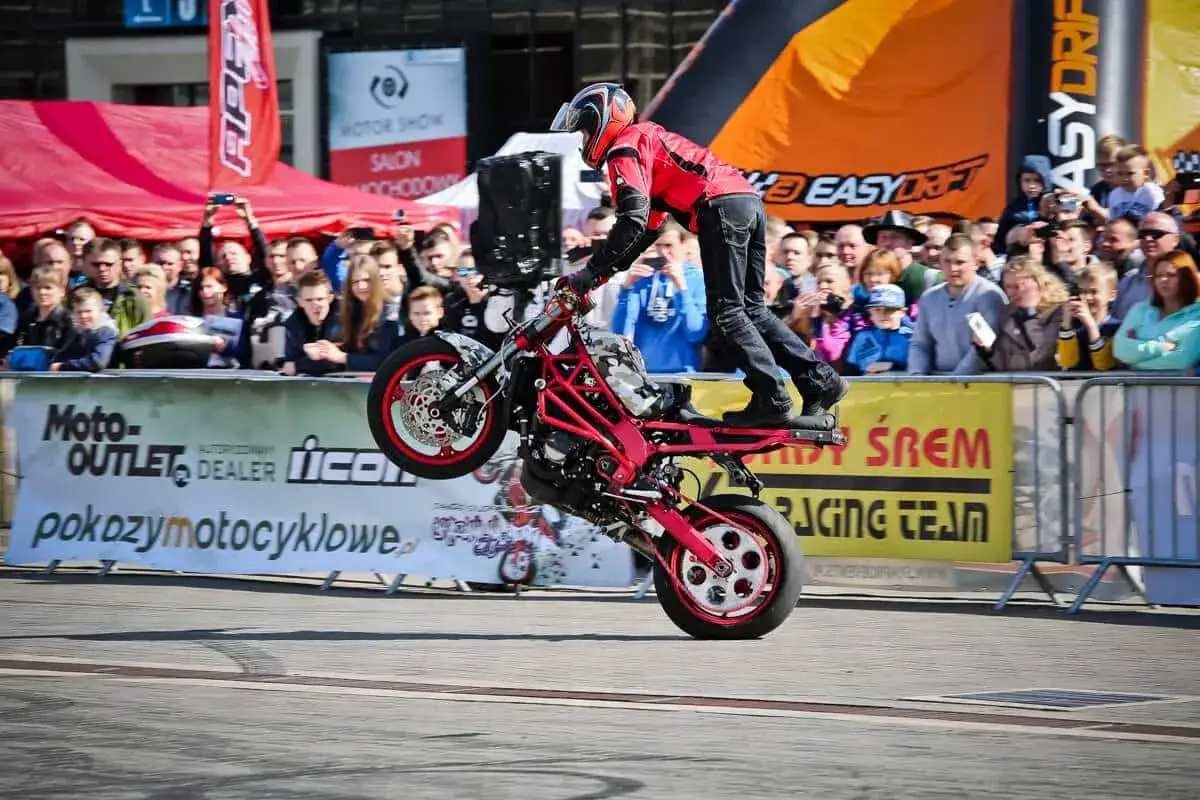 Motocykl do stuntu na tylnym kole, kierowca w czerwonej kurtce, tłum widzów za barierkami.