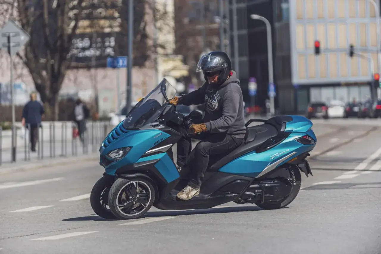 Mężczyzna w kasku jedzie turkusowym skuterem Peugeot Metropolis 400 po mieście.