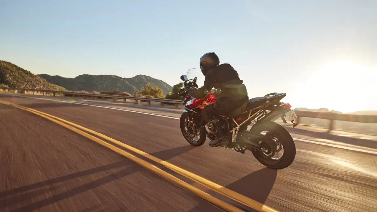 Motocyklista na czerwonym motocyklu Tiger 900 GT Pro przemierza malowniczą górską drogę o zachodzie słońca.