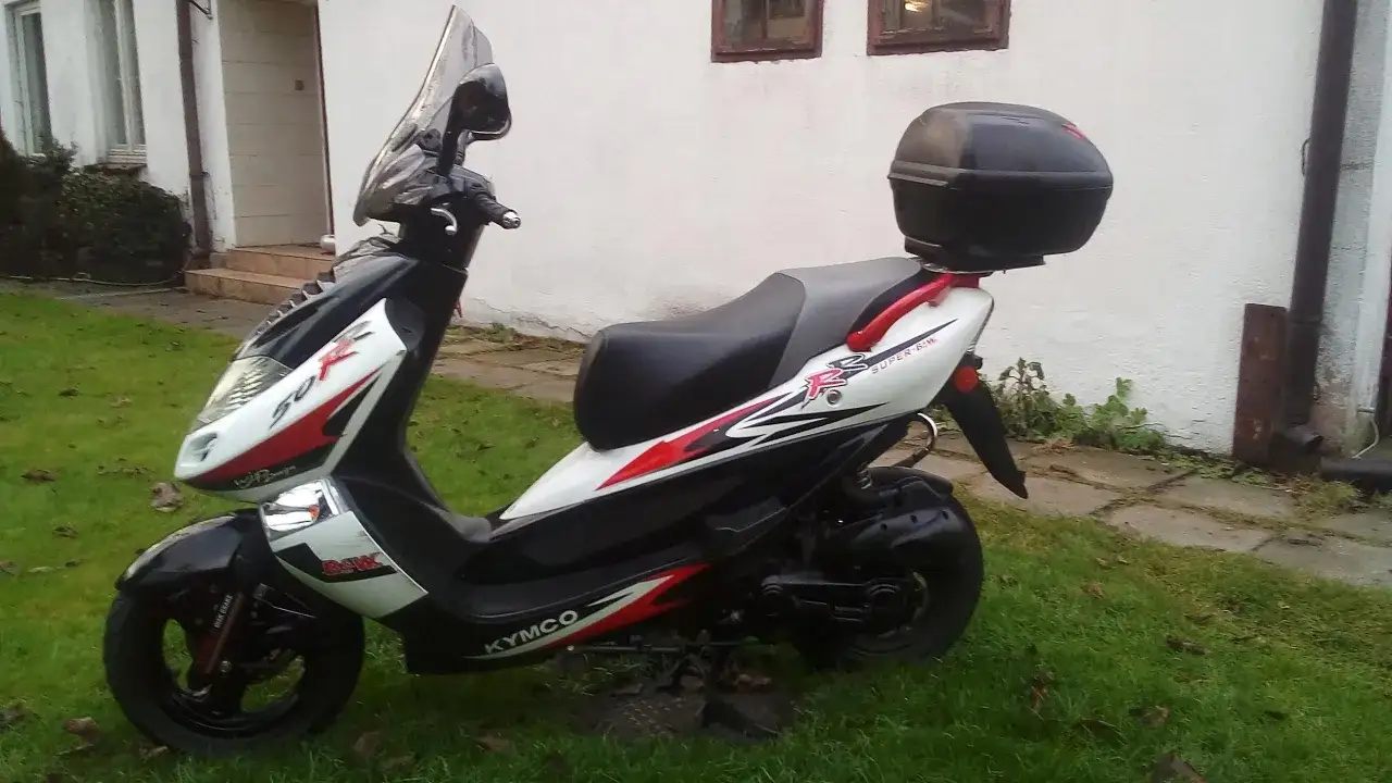 Kymco Bet&Win 50, biało-czarny skuter z czarnym kufrem, zaparkowany na trawie.