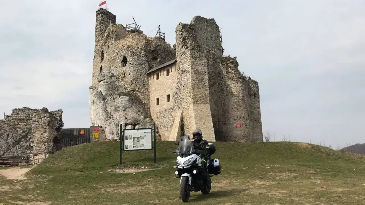Motocyklista na tle ruin zamku na Szlaku Orlich Gniazd. Biały motocykl z kuframi, turysta w kasku i kurtce.