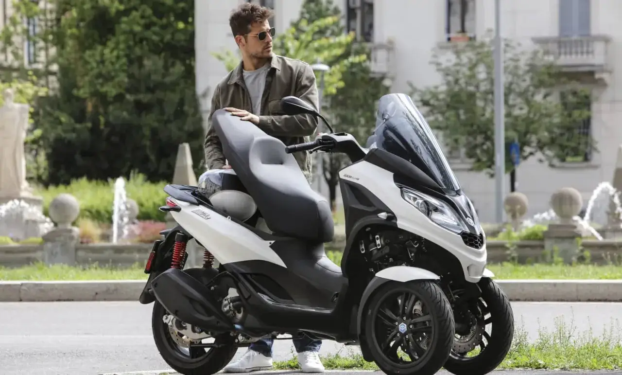 Mężczyzna w okularach przeciwsłonecznych opiera się o biały Piaggio MP3 300. Poznaj jego piaggio mp3 300 dane techniczne.