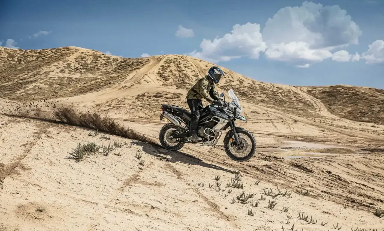 Motocyklista na Triumph Tiger 800 XC wjeżdża na piaszczyste wzgórze, unosząc tumany kurzu. Dane techniczne tego modelu pozwalają na takie wyzwania.