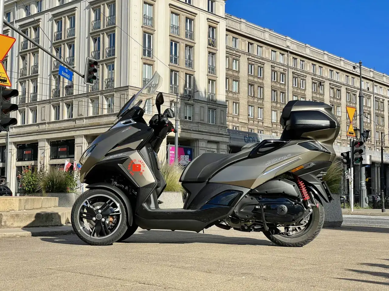 Trójkołowy skuter Peugeot Metropolis 400 na tle zabytkowej kamienicy w słoneczny dzień.