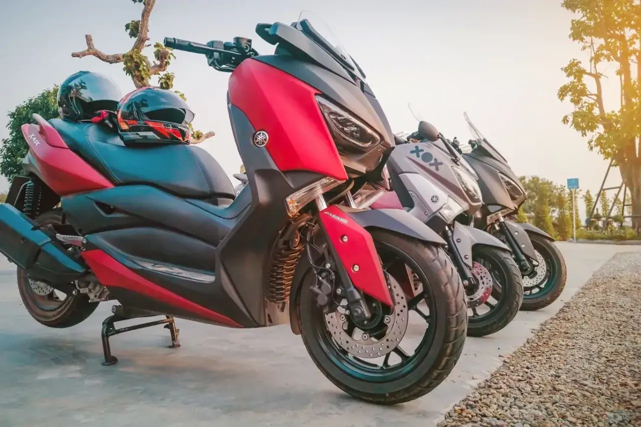 Trzy skutery Yamaha XMAX stoją obok siebie. Czy na skuter trzeba mieć prawo jazdy? Kaski leżą na siedzeniu czerwonego skutera.