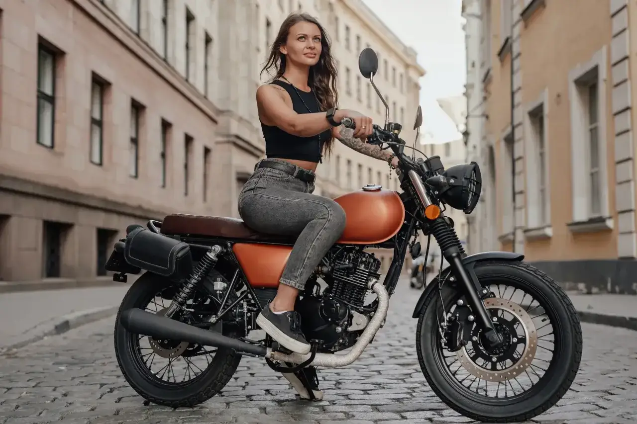 Kobieta na motocyklu 125, w stylowym stroju, cieszy się jazdą po brukowanej uliczce. Docieranie motocykla 125 to dopiero początek przygody.