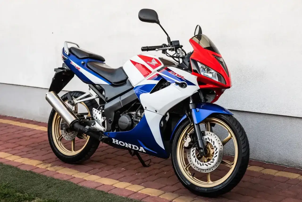 Sportowy motocykl Honda CBR 125R w barwach biało-niebiesko-czerwonych, z złotymi felgami. Idealny na pierwsze wrażenia z jazdy, nie V-max, ale zwinny.