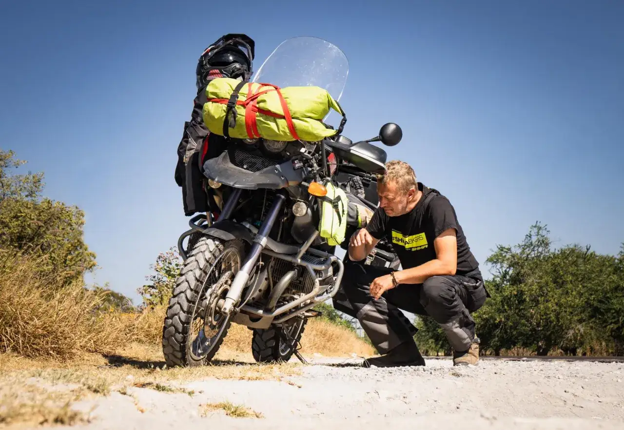 Mężczyzna sprawdza, co zabrać na wyprawę motocyklową. Motocykl załadowany bagażem, kask i zielona torba podr&oacute;żna.