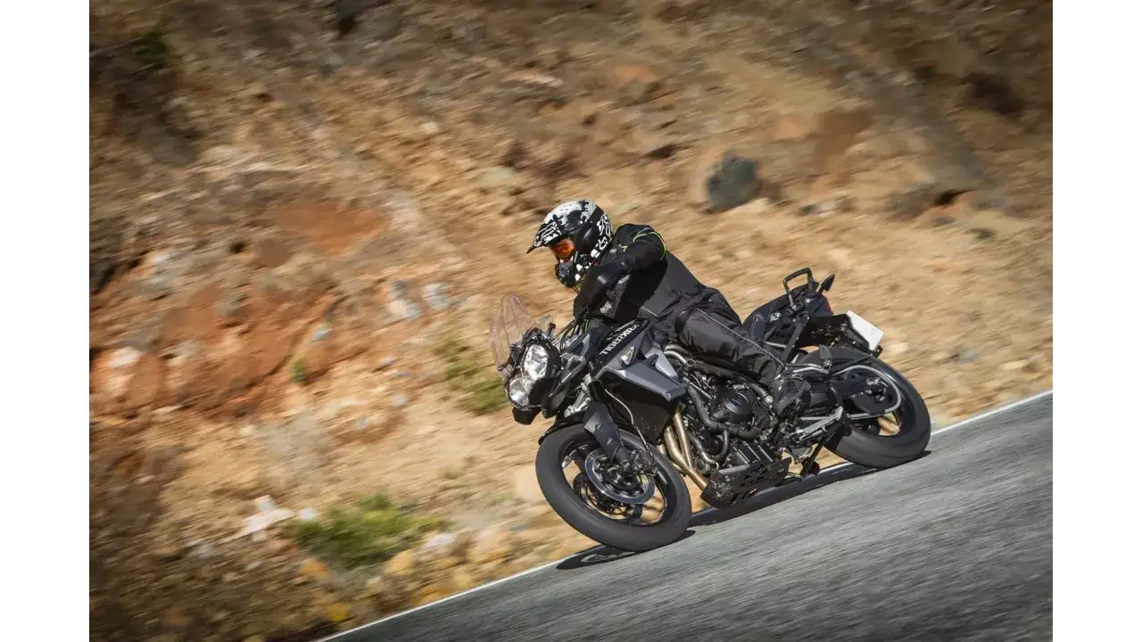 Motocyklista w kasku i odzieży ochronnej pokonuje zakręt na drodze, jadąc motocyklem Triumph Tiger 800 XRX Low.