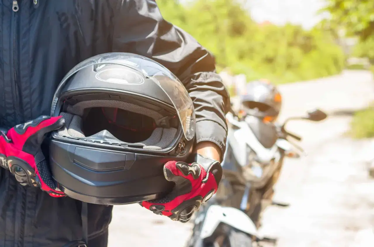 Motocyklista w czerwonych rękawicach trzyma czarny kask. W tle widać motocykl i inne marki kask&oacute;w motocyklowych.