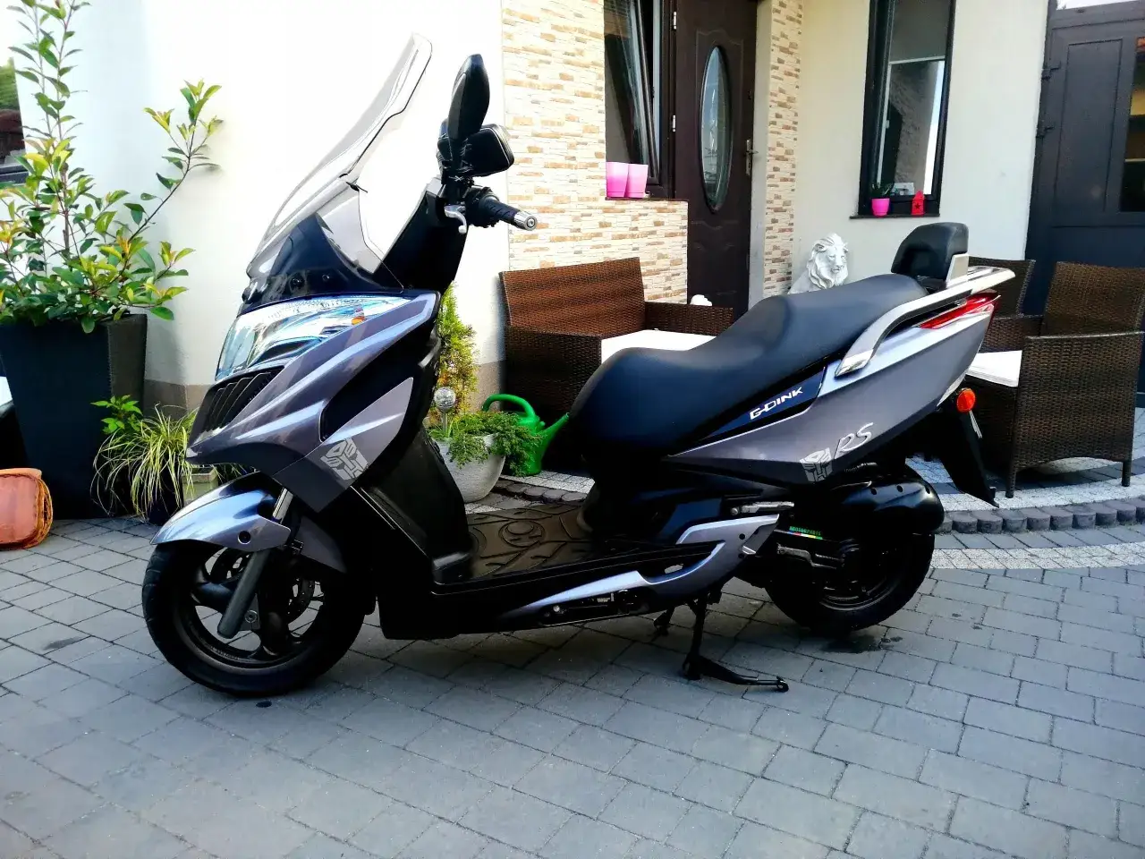 Szary skuter Kymco Dink 50 stoi na podjeździe przed domem, obok roślin i mebli ogrodowych.