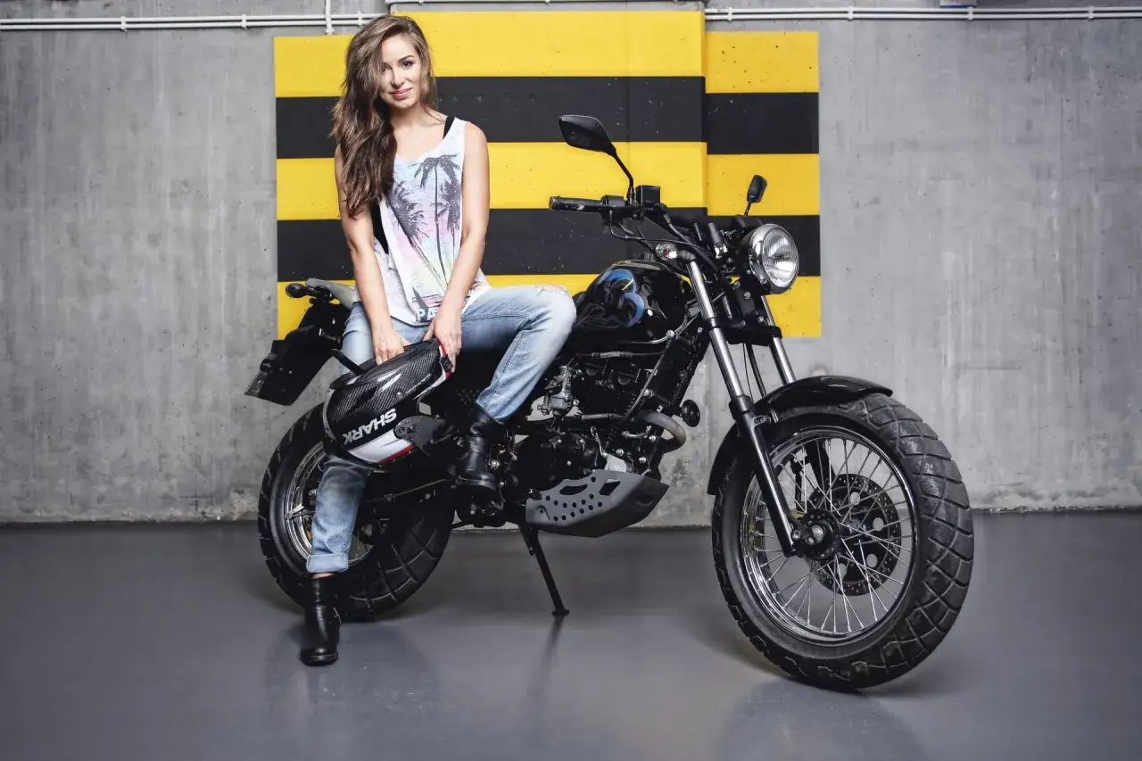 Kobieta w jeansach i koszulce z palmami siedzi na czarnym motocyklu 125, gotowa do docierania. Kask Shark leży obok.