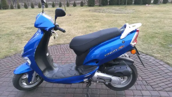 Błękitny skuter Kymco Vitality 50 2T stoi na brukowanym podjeździe, gotowy do jazdy.