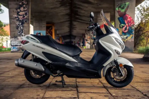 Biały skuter Suzuki Burgman 125, z czarnym siedzeniem, zaparkowany pod mostem z graffiti. Poznaj suzuki burgman 125 dane techniczne.