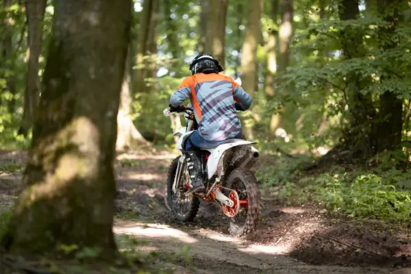 Motocyklista w kasku i pomarańczowej bluzie pędzi leśnym szlakiem. Od ilu lat można jeździć crossem? Odpowiedź zależy od przepisów i umiejętności.