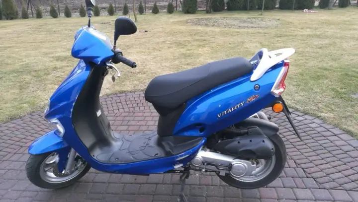 Błękitny skuter Kymco Vitality 50 2T stoi na brukowanym podjeździe, gotowy do jazdy.